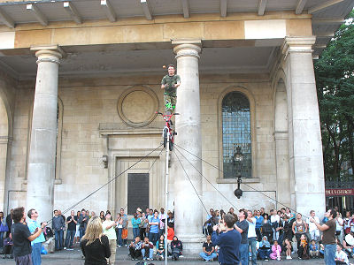 Street performer juggles knives and an apple. Fred is facing away from the camera, holding the rope just to the right of the pole.